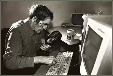 Starszy mężczyzna w okularach z dużą lupą w dłoni patrzy na klawiaturę komputera na której jednym palcem wciska klawisz
